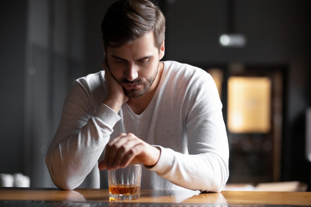 Upset young man in a white shirt is leaning over a drink at a bar. He has his hand on his neck as he looks depressed.