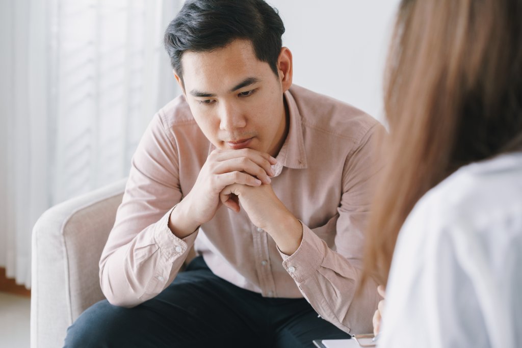 Young Asian man sitting on a therapist's couch with his chin resting on his hands. He's looking down as if in deep thought.