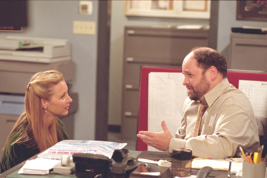 A blonde actress on the left faces a balding actor on the right. They are in mid-conversation and the facial expressions indicate it is serious,