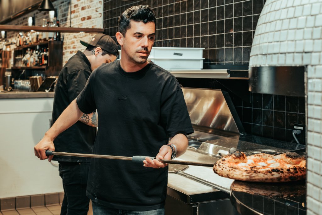 A young dark-haired man in a black t-shirt is putting a pizza on a giant spatula into a pizza oven.