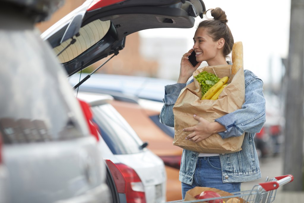 A blonde woman in a denim shirt is talking on the phone while holding a paper bag of groceries next to an open car trunk.