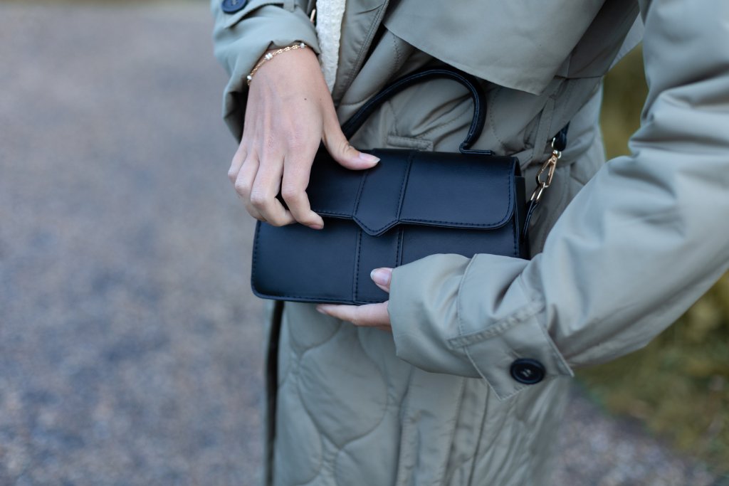 Close up of a woman's torso. She is wearing a gray trenchcoat and reaching in to a crossbody bag on her side.