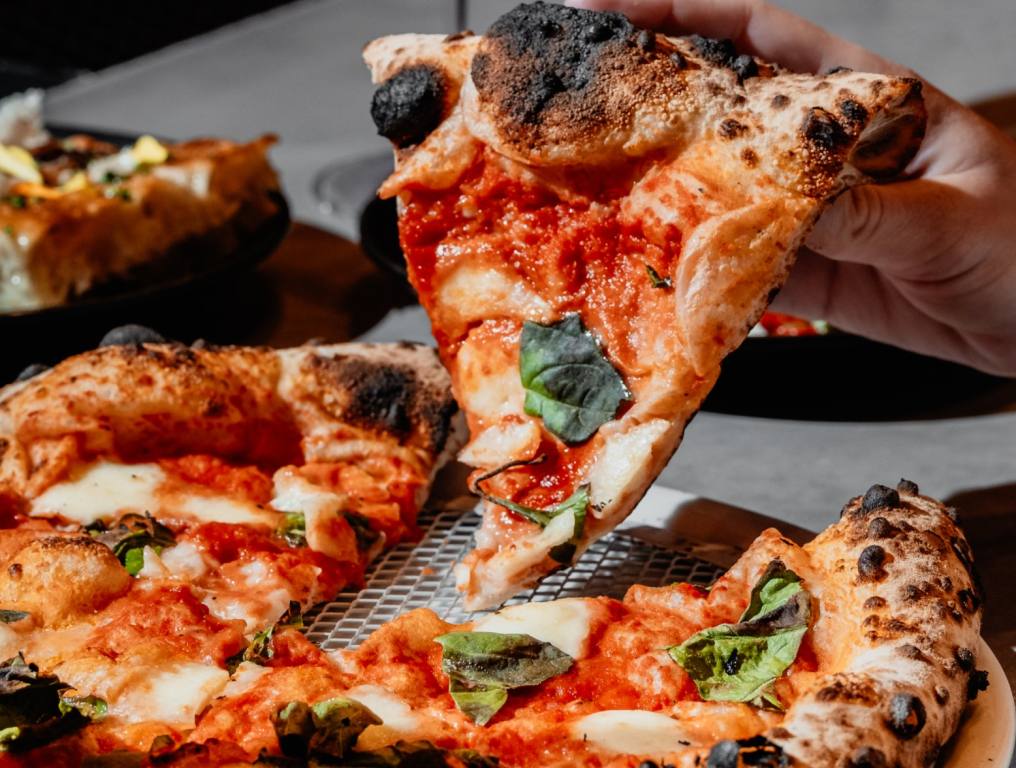 A Margherita pizza from a Vegas pizza shop is shown on a gray table. There is a hand holding up one slice and a wine glass is seen in the background.