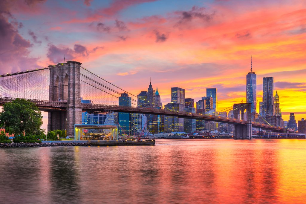 The Brooklyn Bridge stands in front of a Manhattan skyline at dusk with oranges and yellows in the sky.