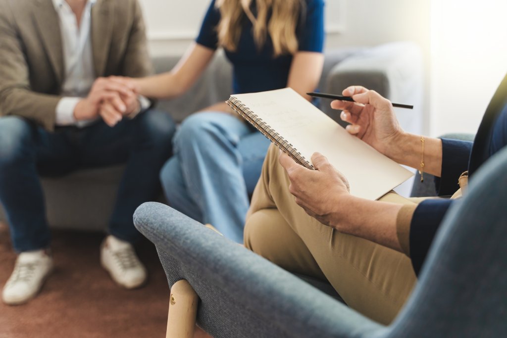 Cropped photo of a couple on a couch in the background with a therapist with a clipboard in a chair in the foreground.