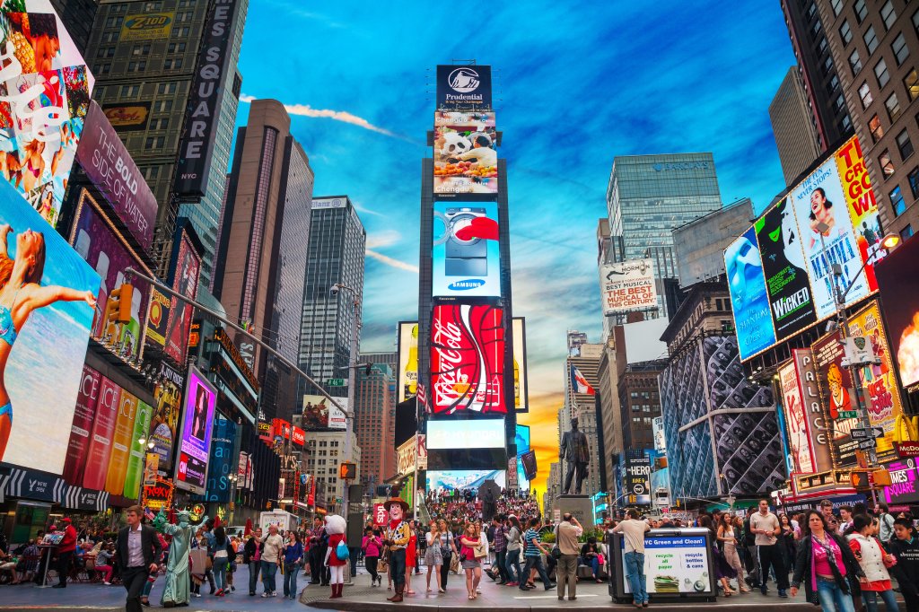 Times Square in New York City is packed with people surrounded by marquis and buildings against a blue sky backdrop.