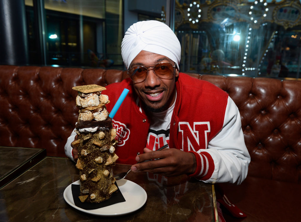 Black actor Nick Cannon sits in a restaurant booth wearing a white head wrap and red sports jacket while enjoying an elaborate s'mores milkshake.