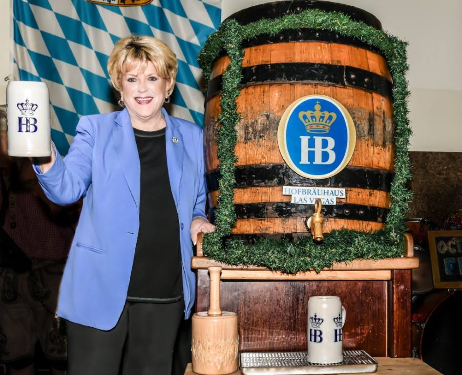 An older woman in a black shirt and pants with a light blue jacket stands next to a large wooden beer keg and smiles.