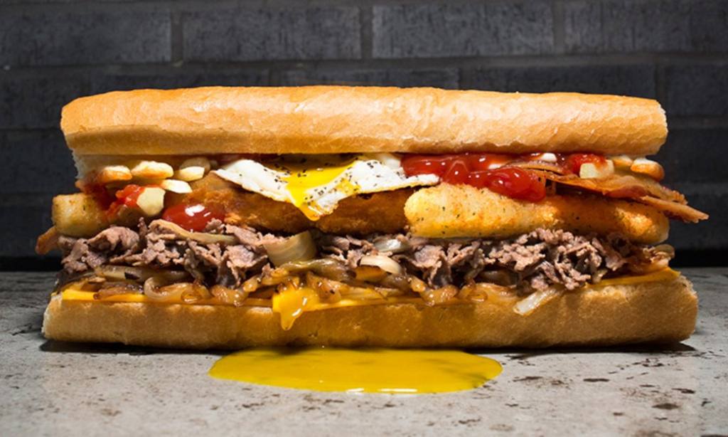 A sandwich sitting on a gray counter is loaded with toppings on a hero roll. There is an egg inside that has broken and the yolk is dripping down the side.