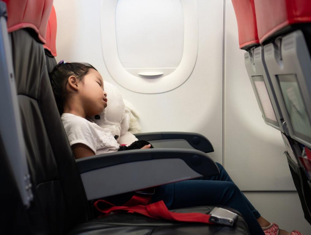 A little girl with dark hair is sleeping on an airplane with her head leaning on the closed window.